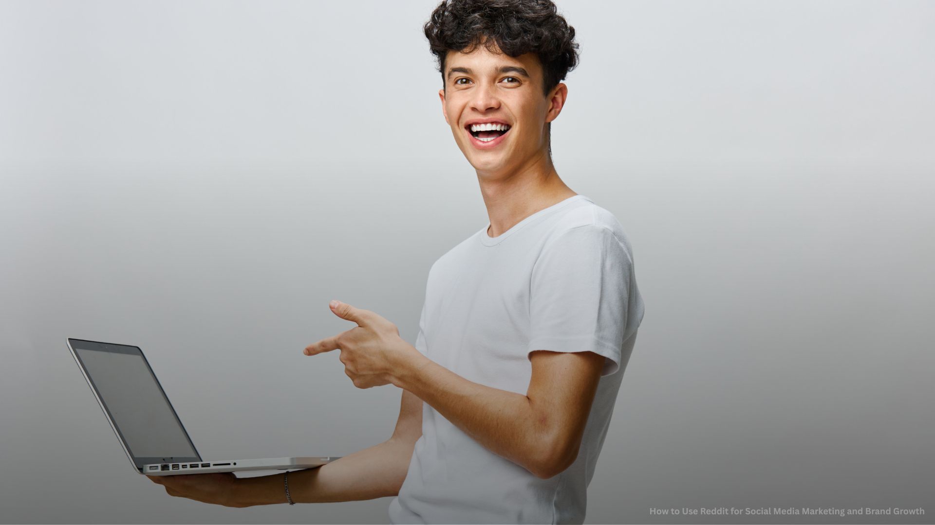 Smiling Young Man with Laptop, Casual Attire with Neutral Gray Background.